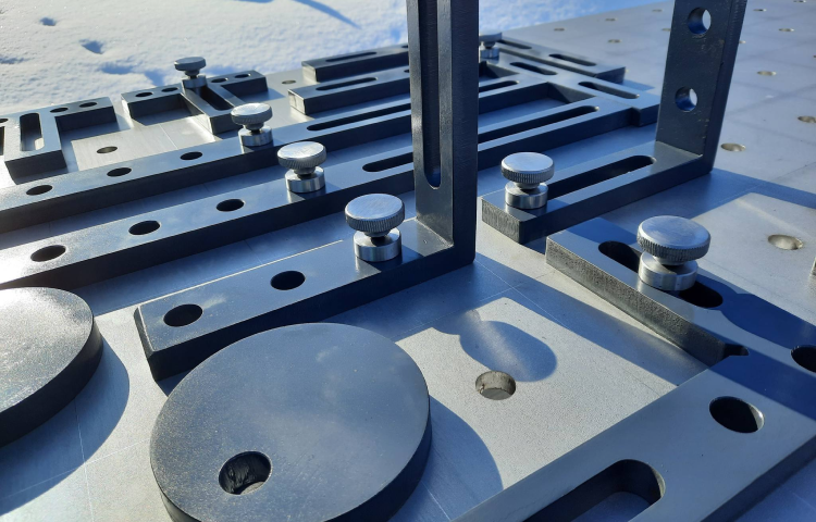 Zbliżenie metalowego przyrządu montażowego z prętami szczelinowymi, okrągłymi płytkami i śrubami zaciskowymi umieszczonymi na perforowanej metalowej powierzchni.