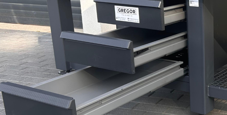 Metalowy schowek z napisem "GREGOR" z otwartymi dwiema dużymi szufladami, umieszczony na utwardzonej powierzchni zewnętrznej.