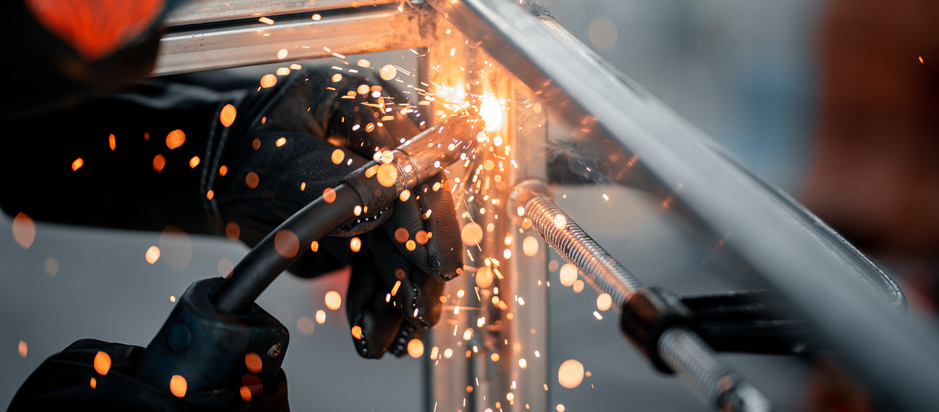 Zbliżenie na osobę w czarnych rękawicach spawającą metal, z jasnopomarańczowymi iskrami unoszącymi się podczas procesu spawania.
