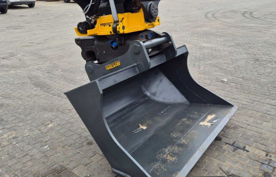 Zbliżenie na żółto-czarny osprzęt koparki z dużą szarą łyżką spoczywającą na utwardzonej powierzchni.