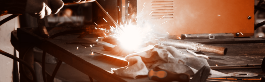 Osoba spawa metalowe elementy na stole warsztatowym, wytwarzając jasne iskry. W pobliżu widoczne są różne narzędzia i ściereczka.