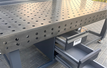 Metalowy stół warsztatowy z perforowanym blatem i dwiema otwartymi szufladami, stojący na utwardzonej powierzchni.