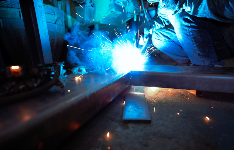 Osoba spawa metalowe belki w warsztacie, tworząc jasne niebieskie iskry i światło.